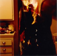 A man stands in the kitchen as he bursts into flame. He doesn't seem to be feeling any pain. Perhaps this has to do with all the medication bottles found along the counter. His eyes look at the viewer and sparkle from deep empty sockets. Flames erupt around him and burst from inside him. This is a scary image.